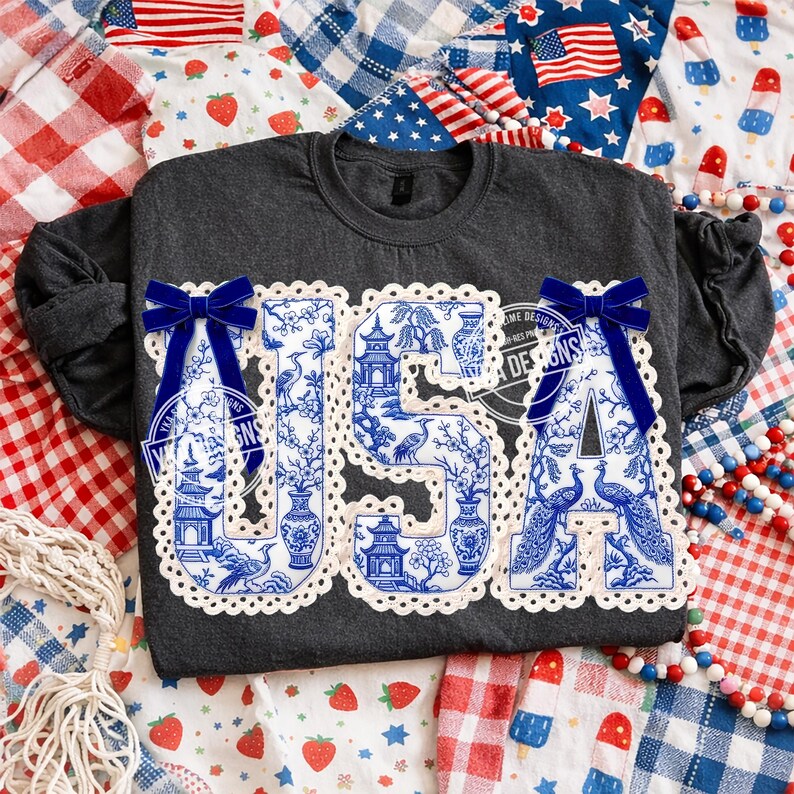 May include: A dark grey sweatshirt featuring the word "USA" in a blue and white chinoiserie pattern. The letters are edged with white lace and have blue velvet bows. The background includes patriotic fabrics.