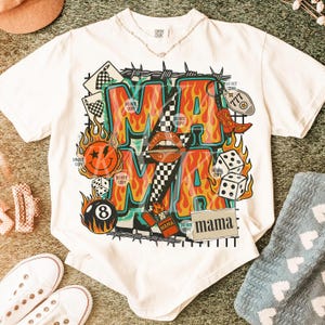 Puede incluir: Camiseta blanca con un dise&ntilde;o gr&aacute;fico audaz. El dise&ntilde;o presenta la palabra "MAMA" en llamas naranjas, rodeada de dados, una carita sonriente y una bota de vaquero. La camiseta tambi&eacute;n tiene el texto "DO NOT COPY".