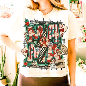 May include: Cream-coloured t-shirt featuring a festive "MAMA" graphic. The design includes Santa Claus, candy canes, playing cards, and a lucky 777 charm. The words "DO NOT COPY" are also visible.