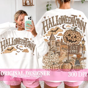May include: A white long-sleeve sweatshirt featuring a leopard print graphic of the text HALLOWEENTOWN, a jack-o'-lantern, a ghost, a haunted house, bats, and a sign that reads EST. 1998. The design includes stars, clouds, and a scarecrow.