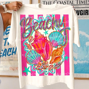 Op de afbeelding: Wit T-shirt met een kleurrijk strandthema. Het ontwerp toont het woord "Beachy" in teal en gouden glitter, samen met illustraties van schelpen in roze, oranje en teal. Roze en witte strepen omlijsten het ontwerp.