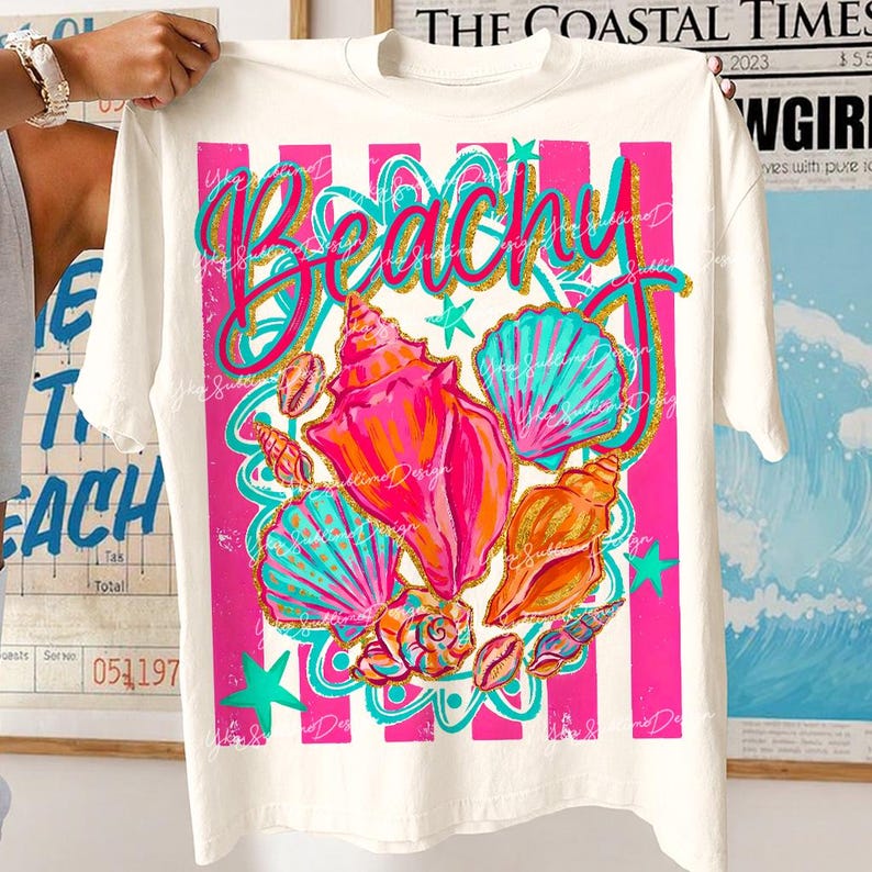 Op de afbeelding: Wit T-shirt met een kleurrijk strandthema. Het ontwerp toont het woord "Beachy" in teal en gouden glitter, samen met illustraties van schelpen in roze, oranje en teal. Roze en witte strepen omlijsten het ontwerp.
