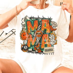 Puede incluir: Camiseta blanca con un dise&ntilde;o gr&aacute;fico colorido. El dise&ntilde;o presenta la palabra "MAMA" en letras de fuego, rodeada de elementos como naipes, dados y una carita sonriente. La camiseta tiene un cuello redondo cl&aacute;sico.