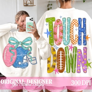 Puede incluir: Una sudadera blanca de manga larga con un gráfico de un balón de fútbol americano rosa y un casco azul con un lazo turquesa en el frente, y la palabra TOUCHDOWN en letras coloridas y estampadas con motivos de fútbol y estrellas en la espalda.