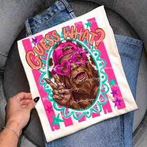 May include: Off-white t-shirt with a graphic of a Bigfoot wearing a pink cap and heart-shaped sunglasses, giving a peace sign. The text "GUESS WHAT?" is in orange and pink. The design includes pink stripes, teal and purple stars, and a decorative border.