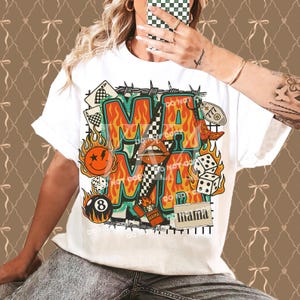Puede incluir: Camiseta blanca con un dise&ntilde;o gr&aacute;fico colorido. El dise&ntilde;o presenta la palabra "MA" en letras grandes, rodeada de llamas e ilustraciones retro. Incluye dados, una carita sonriente y la palabra "mama".