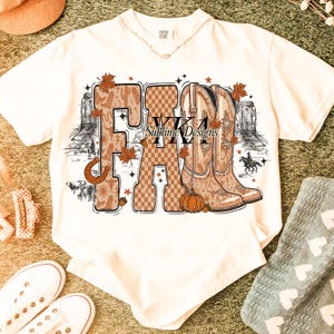 May include: Cream-coloured t-shirt with a fall-themed graphic. The word "FALL" is displayed with a horseshoe, cowboy boots, and autumn leaves. The graphic includes a pumpkin and the text "WKA Sublime Designs."