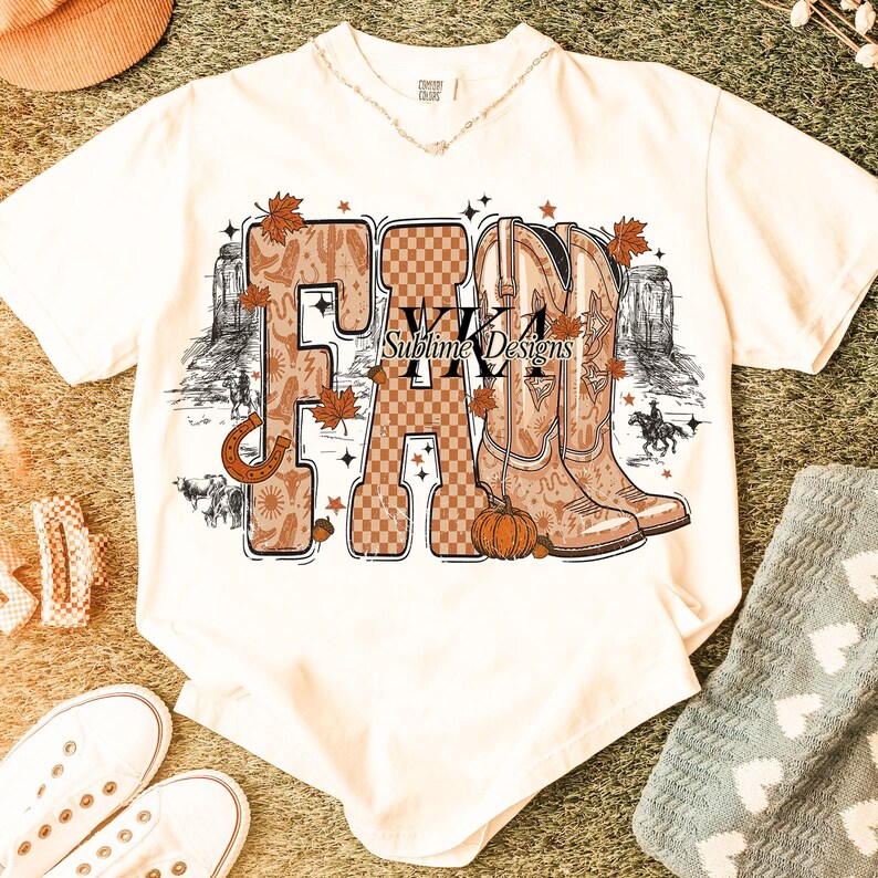May include: Cream-coloured t-shirt with a fall-themed graphic. The word "FALL" is displayed with a horseshoe, cowboy boots, and autumn leaves. The graphic includes a pumpkin and the text "WKA Sublime Designs."