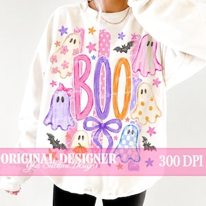 May include: A cream-colored long-sleeve sweatshirt featuring a graphic print with the text BOO, colorful ghosts, bats, stars, and a bow. The ghosts have various patterns including flowers, polka dots, and checkers in pink, purple, and orange hues.