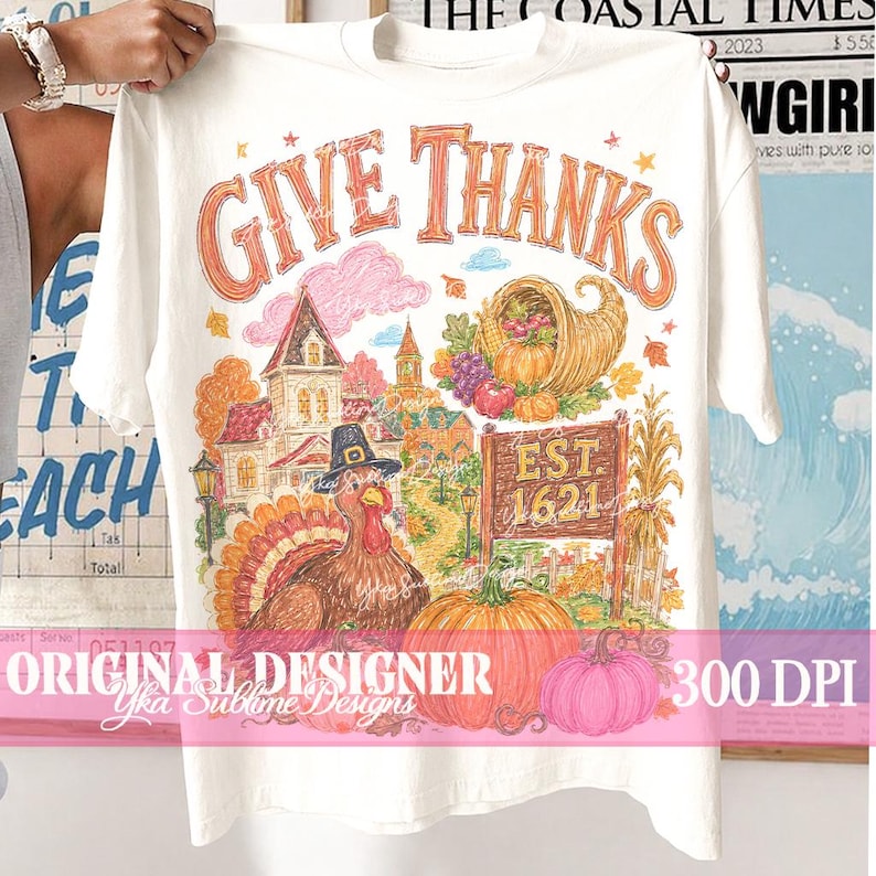 May include: A cream-colored t-shirt featuring a colorful graphic print with the text GIVE THANKS and EST. 1621. The illustration includes a turkey wearing a pilgrim hat, a cornucopia, pumpkins, a house, and autumn foliage in shades of orange and brown.