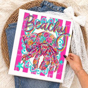 May include: A white canvas print with a vibrant design featuring a floral-patterned crab and the word "Beachy". The crab is in shades of pink, orange, and teal, set against a pink and white striped background. The print is held by a person.