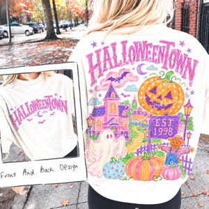 May include: A cream-colored t-shirt featuring a graphic design with the text HALLOWEENTOWN in pink. The back design displays a colorful illustration of a haunted house, a large jack-o'-lantern, a ghost, a scarecrow, and a sign reading EST. 1998.