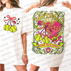 Puede incluir: Camiseta blanca con dise&ntilde;o de softbol. El gr&aacute;fico incluye dos pelotas de softbol con lazos, un coraz&oacute;n, bates y el texto "SOFTBALL HOME RUN HERO". El dise&ntilde;o utiliza colores rosa, verde y amarillo.