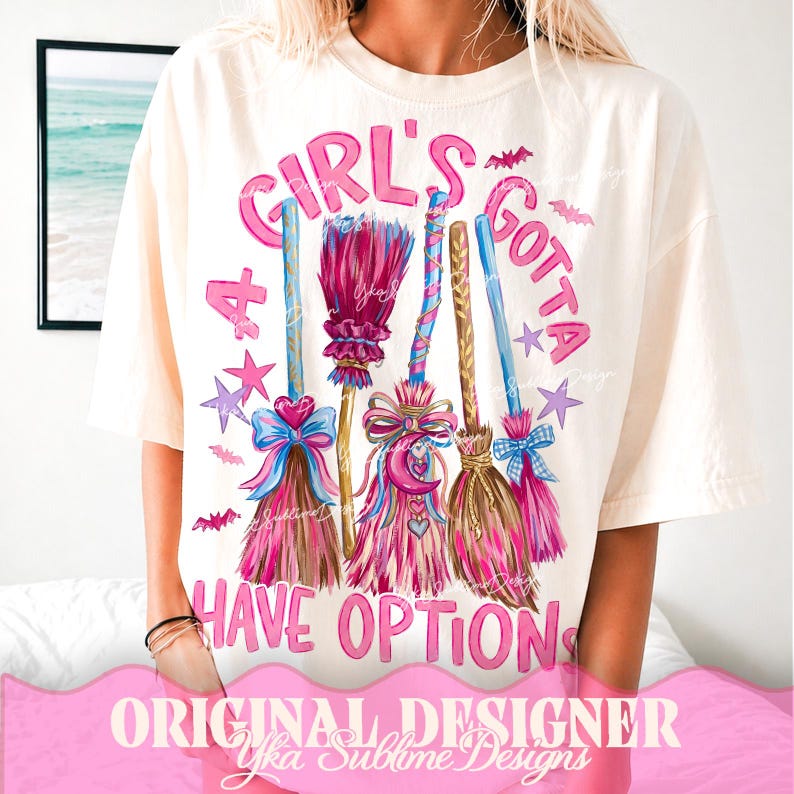 May include: A cream-colored short-sleeve t-shirt featuring a graphic print of five colorful broomsticks with bows and stars. The text A GIRL'S GOTTA HAVE OPTIONS is printed in pink around the brooms, accented by small purple stars and bats.