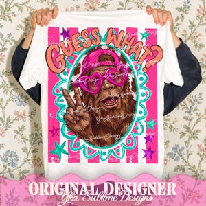 May include: White t-shirt featuring a cartoon Bigfoot wearing pink heart-shaped sunglasses and a backwards cap. The shirt has the text "GUESS WHAT?" in a bubble font, with pink and teal accents. The design includes stars and a decorative border.