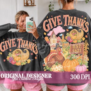 May include: A charcoal gray crewneck sweatshirt featuring a graphic print with the text GIVE THANKS. The design includes a cornucopia, a turkey wearing a hat, a house, and pumpkins in orange, pink, and green tones. The back shows the full design.