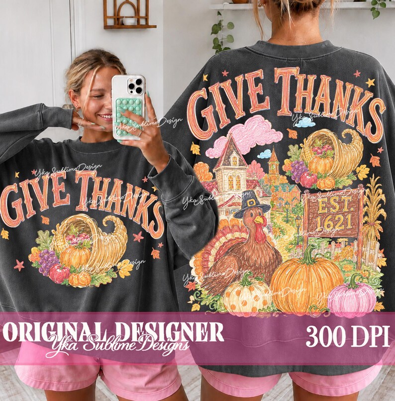 May include: A charcoal gray crewneck sweatshirt featuring a graphic print with the text GIVE THANKS. The design includes a cornucopia, a turkey wearing a hat, a house, and pumpkins in orange, pink, and green tones. The back shows the full design.