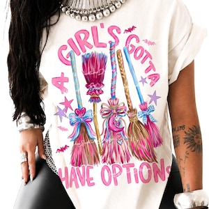 May include: An off-white t-shirt featuring a graphic print of four colorful broomsticks with pink, blue, and brown bristles. The text "A GIRL'S GOTTA HAVE OPTIONS" is printed in pink letters around the brooms, accented by small pink bats and stars.