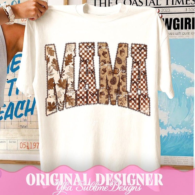 May include: A white short-sleeve t-shirt featuring the word MAMA printed in large block letters. The letters are filled with a repeating pattern of brown autumn leaves, checkered squares, acorns, and pinecones, all outlined with a scalloped border.