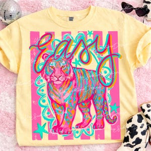 May include: A light yellow t-shirt with a vibrant graphic of a tiger and the word "easy" in a colorful, playful font. The tiger is depicted with pink, blue, and orange stripes and spots, set against a pink and white striped background.