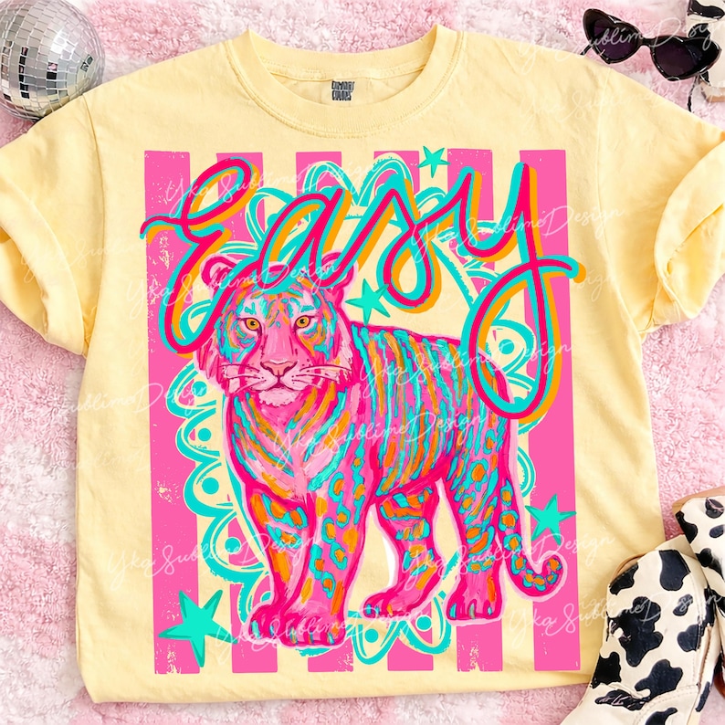 May include: A light yellow t-shirt with a vibrant graphic of a tiger and the word "easy" in a colorful, playful font. The tiger is depicted with pink, blue, and orange stripes and spots, set against a pink and white striped background.