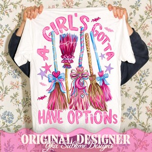 May include: A white t-shirt featuring a graphic print of four stylized brooms in shades of pink, blue, and brown. The text "A GIRL'S GOTTA HAVE OPTIONS" is printed in pink letters, accompanied by small purple stars and pink bat illustrations.