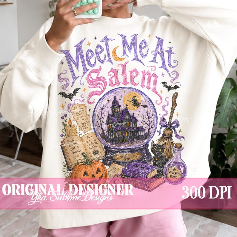 May include: A cream-coloured sweatshirt featuring a graphic print with the text "Meet Me At Salem" in purple and pink. The design includes a crystal ball with a haunted house, a witch on a broom, a pumpkin, an open spellbook, a broom, and a cat.