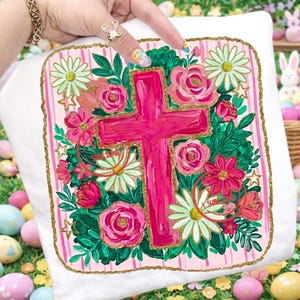 Peut inclure: Coussin blanc avec un motif de croix rose entour&eacute; de fleurs. La croix est peinte en rose vif, et les fleurs comprennent des roses roses, des marguerites et d'autres fleurs. Le coussin a une bordure dor&eacute;e scintillante et des rayures roses.