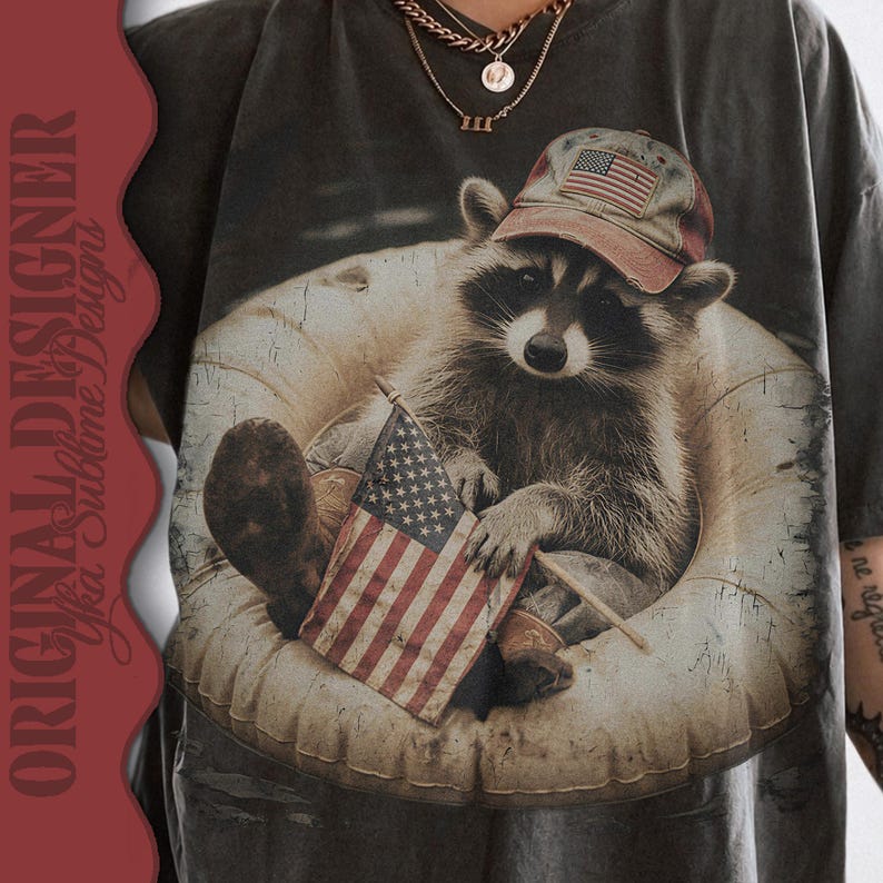 Puede incluir: Camiseta gris oscuro con un mapache que lleva un sombrero con la bandera estadounidense, sosteniendo una peque&ntilde;a bandera y sentado en una c&aacute;mara de aire. La camiseta tiene un aspecto vintage y desgastado.