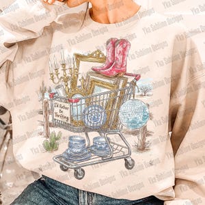 May include: A beige sweatshirt featuring a shopping cart filled with vintage items. The cart holds pink cowboy boots, picture frames, and blue and white china. A sign reads "I'd Rather Be Thrifting."