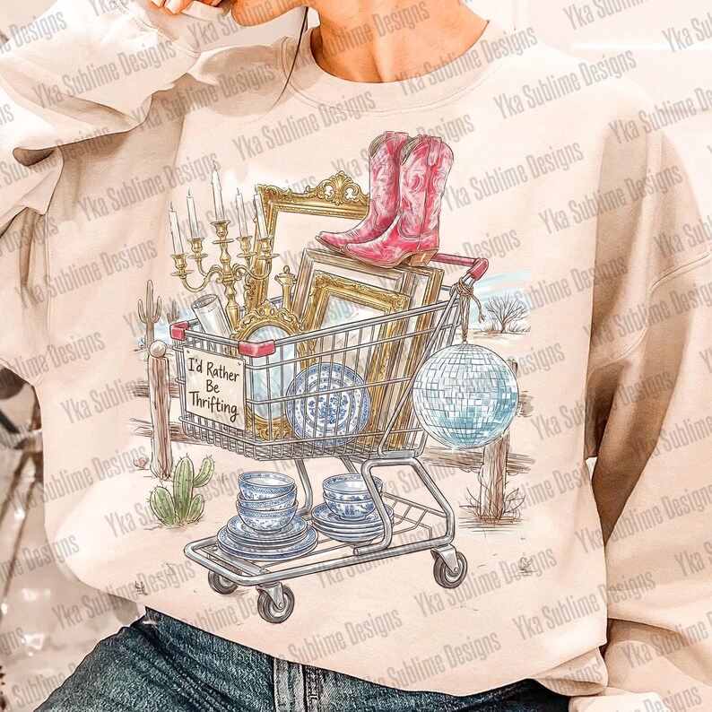 May include: A beige sweatshirt featuring a shopping cart filled with vintage items. The cart holds pink cowboy boots, picture frames, and blue and white china. A sign reads "I'd Rather Be Thrifting."
