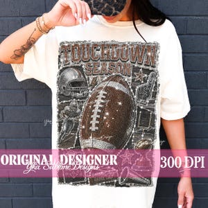 May include: An off-white t-shirt featuring a graphic print with the text TOUCHDOWN SEASON. The design includes a brown football, a silver helmet, a goal post, a trophy, a megaphone, and stars, all rendered with a sparkling, glittery texture.