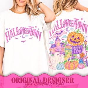 May include: A white t-shirt featuring a pink graphic on the front that reads HALLOWEENTOWN with stars, a moon, and bats. The back displays a colorful illustration of a haunted house, a large jack-o'-lantern, a ghost, and a sign reading EST. 1998.