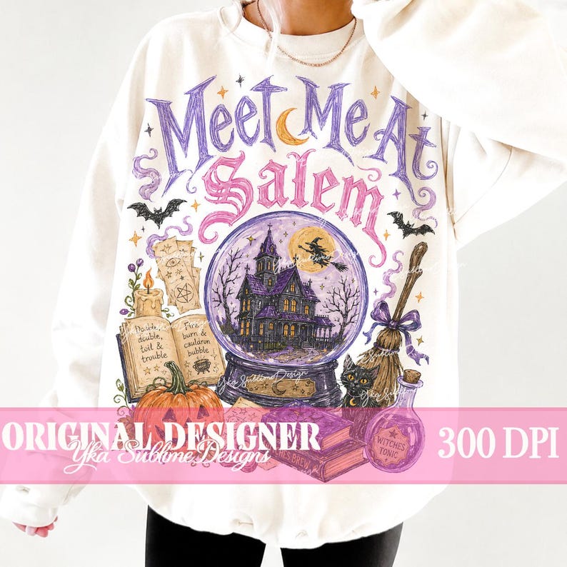 May include: A cream-coloured sweatshirt featuring a graphic print with the text "Meet Me At Salem" in purple and pink. The design includes a central crystal ball with a haunted house, a witch on a broom, a pumpkin, an open spellbook, a cat, and a potion.