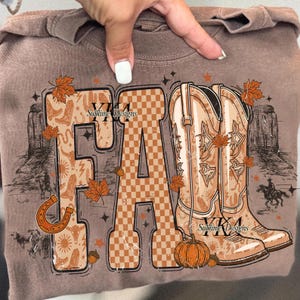May include: Brown t-shirt with a graphic design featuring the word "FALL" in a mix of patterns and fonts. The design includes cowboy boots, autumn leaves, a horseshoe, and a pumpkin. The text "Sublime Designs" is also visible.