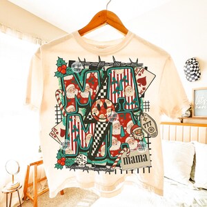 May include: A cream-coloured t-shirt with a graphic design featuring the word "MAMA" in various fonts and patterns. The design includes Santa Claus faces, candy canes, playing cards, and a smiley face. The shirt is hanging on a wooden hanger.