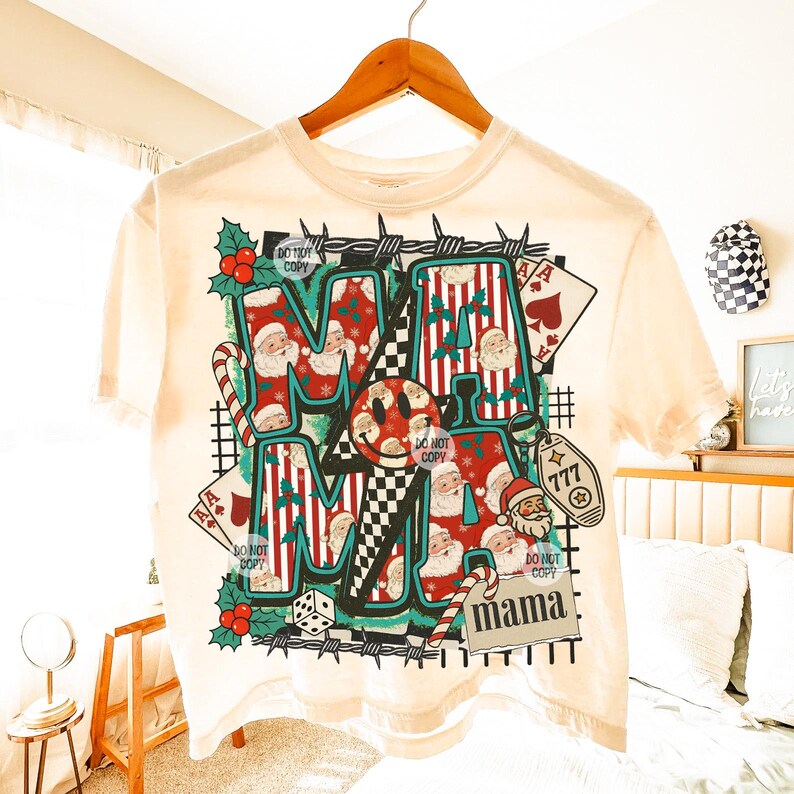 May include: A cream-coloured t-shirt with a graphic design featuring the word "MAMA" in various fonts and patterns. The design includes Santa Claus faces, candy canes, playing cards, and a smiley face. The shirt is hanging on a wooden hanger.