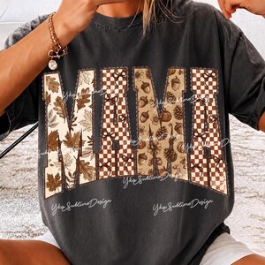 May include: A charcoal gray short-sleeve t-shirt featuring the word MAMA in large block letters. The letters are filled with a pattern of brown autumn leaves, a brown and white checkerboard design, and brown acorns and pinecones on a tan background.