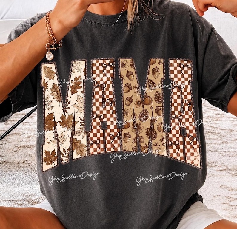 May include: A charcoal gray short-sleeve t-shirt featuring the word MAMA in large block letters. The letters are filled with a pattern of brown autumn leaves, a brown and white checkerboard design, and brown acorns and pinecones on a tan background.