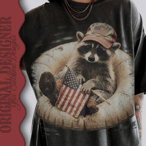 Puede incluir: Camiseta gris oscuro con un mapache que lleva una gorra de b&eacute;isbol con la bandera estadounidense. El mapache est&aacute; sentado en una c&aacute;mara de aire y sostiene una peque&ntilde;a bandera estadounidense. La camiseta tiene un aspecto vintage y desgastado.