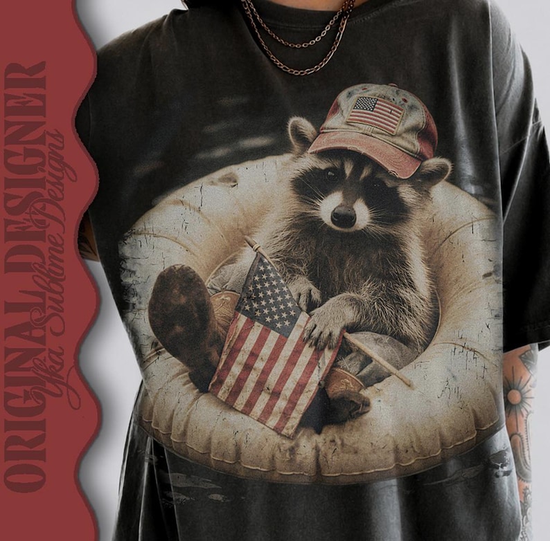 Puede incluir: Camiseta gris oscuro con un mapache que lleva una gorra de b&eacute;isbol con la bandera estadounidense. El mapache est&aacute; sentado en una c&aacute;mara de aire y sostiene una peque&ntilde;a bandera estadounidense. La camiseta tiene un aspecto vintage y desgastado.