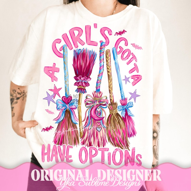 May include: A white t-shirt featuring a graphic print of five stylized broomsticks in shades of pink, blue, and brown, decorated with bows and stars. The text reads A GIRL'S GOTTA HAVE OPTIONS in pink lettering, surrounded by small pink bats and stars.