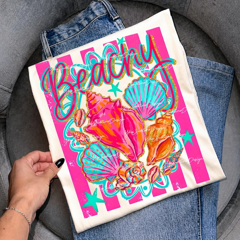 Op de afbeelding: Een wit T-shirt met een strandthema. Het shirt heeft het woord "Beachy" in een cursief lettertype, samen met kleurrijke schelpen in roze, oranje en turquoise. Roze strepen en turquoise sterren maken het ontwerp compleet.