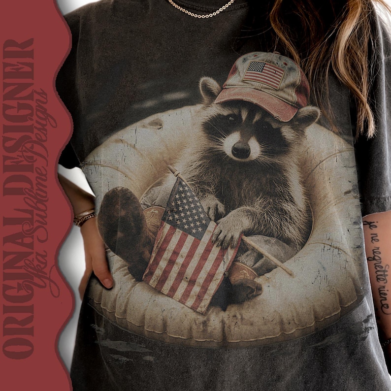 Puede incluir: Camiseta gris oscuro con un mapache que lleva una gorra de b&eacute;isbol con una bandera estadounidense. El mapache sostiene una peque&ntilde;a bandera estadounidense y est&aacute; sentado en un flotador inflable redondo. La camiseta tiene el texto "ORIGINAL DESIGNER Yfka Sublime Designs".