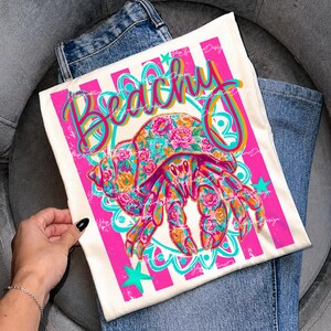 May include: Off-white t-shirt with a colorful graphic of a hermit crab adorned with floral patterns. The word "Beachy" is written in a vibrant, cursive font, framed by pink stripes and teal accents. The shirt is laid on top of blue jeans.