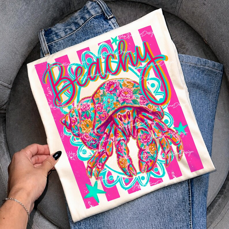 May include: Off-white t-shirt with a colorful graphic of a hermit crab adorned with floral patterns. The word "Beachy" is written in a vibrant, cursive font, framed by pink stripes and teal accents. The shirt is laid on top of blue jeans.