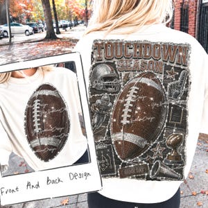 May include: A cream-colored t-shirt featuring a brown football graphic on the front and a large "TOUCHDOWN SEASON" graphic on the back. The back design includes a football, helmet, player, trophy, megaphone, and play diagram with glitter-like accents.