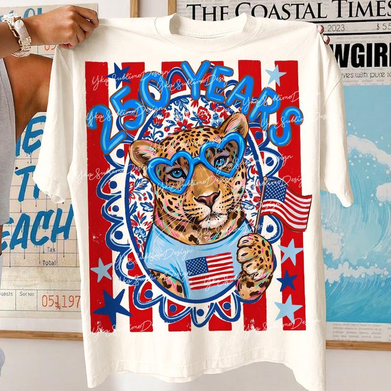 Op de afbeelding: Wit T-shirt met een patriottisch ontwerp. De afbeelding toont een luipaard met een hartvormige bril en een Amerikaanse vlag. De tekst "250 YEARS" is blauw, met rode en blauwe sterren en strepen.