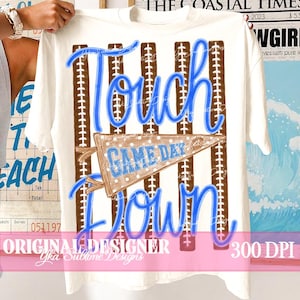 May include: A white t-shirt featuring a graphic design with vertical brown football-textured stripes. The text "Touch Down" is written in a bright blue script font, and a brown pennant with white polka dots displays the text "GAME DAY".