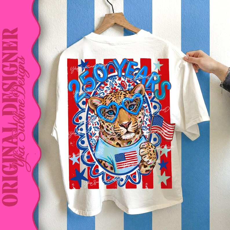 Op de afbeelding: Wit T-shirt met een luipaard met een blauwe hartvormige bril en een Amerikaanse vlag. Het shirt heeft een rood, wit en blauw ontwerp met de tekst "250 YEARS". Het T-shirt hangt aan een houten hanger.